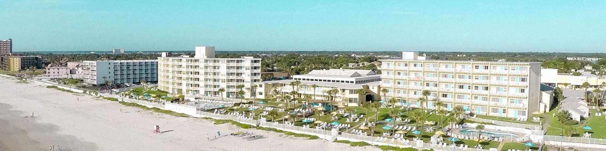 The image depicts a coastal scene with a sandy beach lined with several large buildings or hotels, stretching along the shoreline.