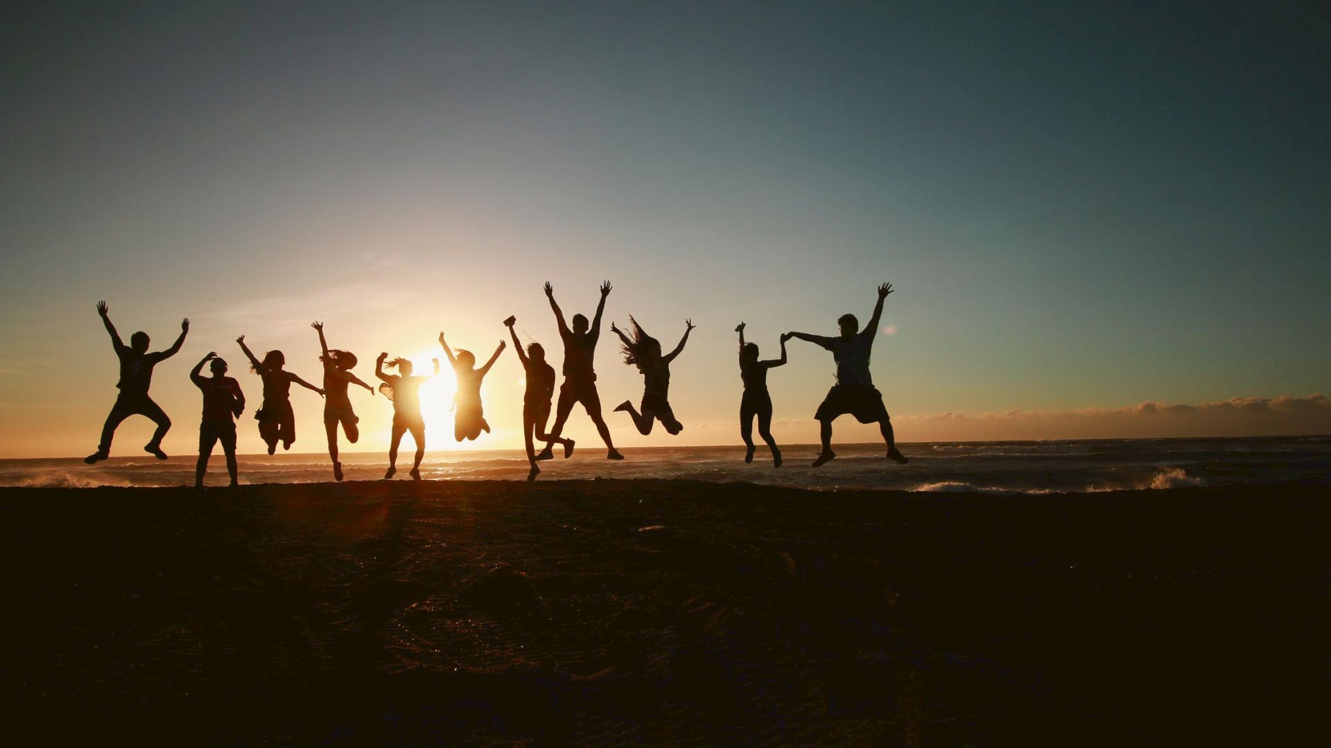 A group of people jumps in unison on a beach, silhouetted against a sunset, creating a lively and joyful scene.