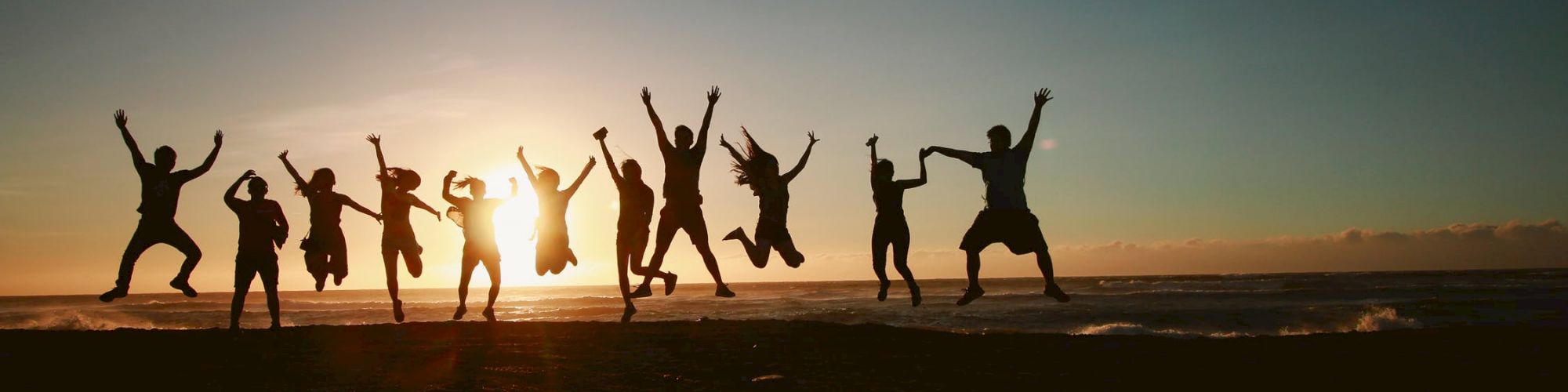 A group of people jumps in unison on a beach, silhouetted against a sunset, creating a lively and joyful scene.