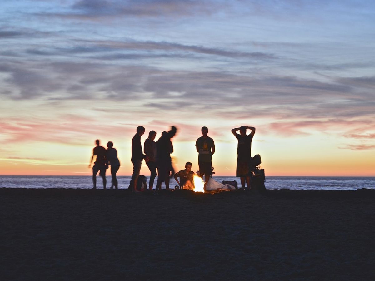 A group of people is gathered around a campfire on a beach during sunset, creating silhouetted figures against the colorful sky.