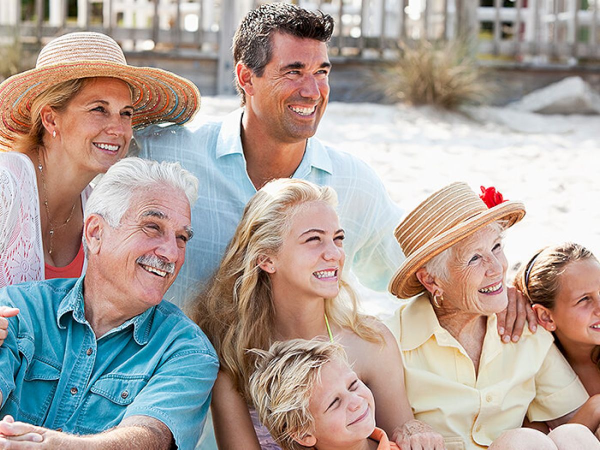 A group of seven people, of various ages, are smiling and sitting together on a beach with sandy surroundings.