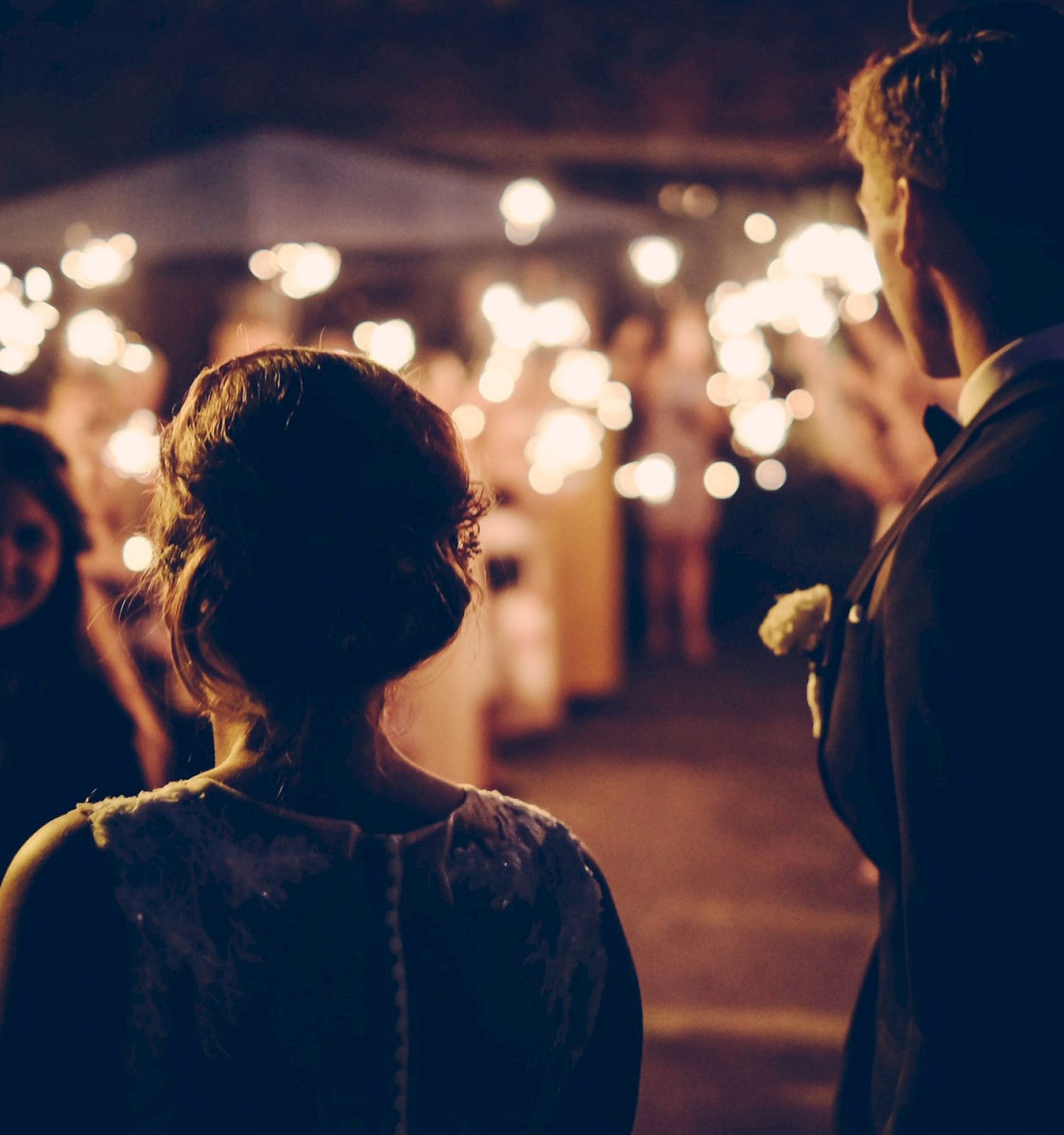 A couple stands facing a crowd holding sparklers during a nighttime celebration or event, creating a festive and romantic atmosphere.