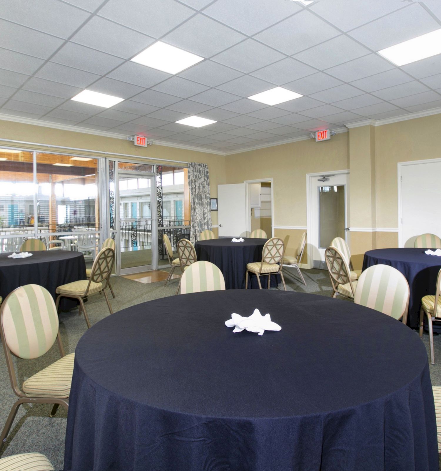 The image shows a banquet room with round tables covered in navy-blue tablecloths, surrounded by chairs. Large windows are in the background.