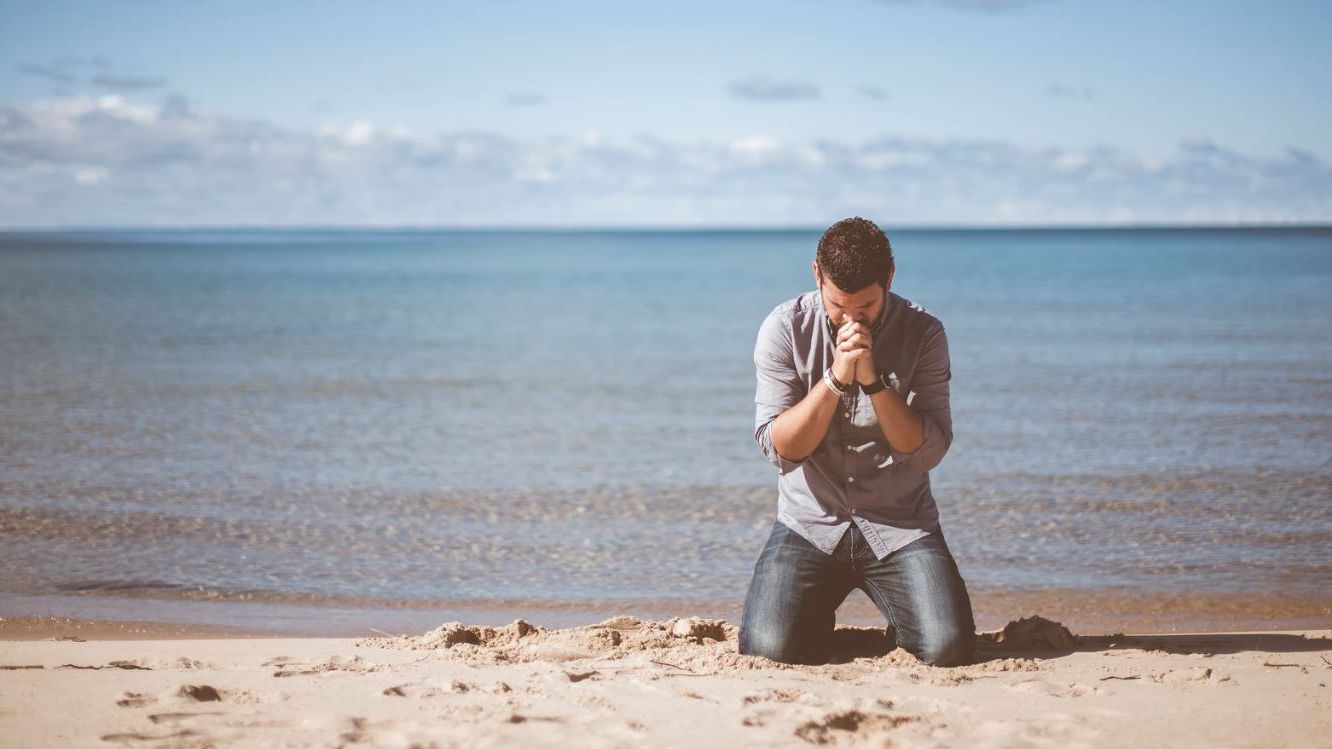 A person is kneeling on a sandy beach, clasping their hands together, facing the ocean under a partly cloudy sky.