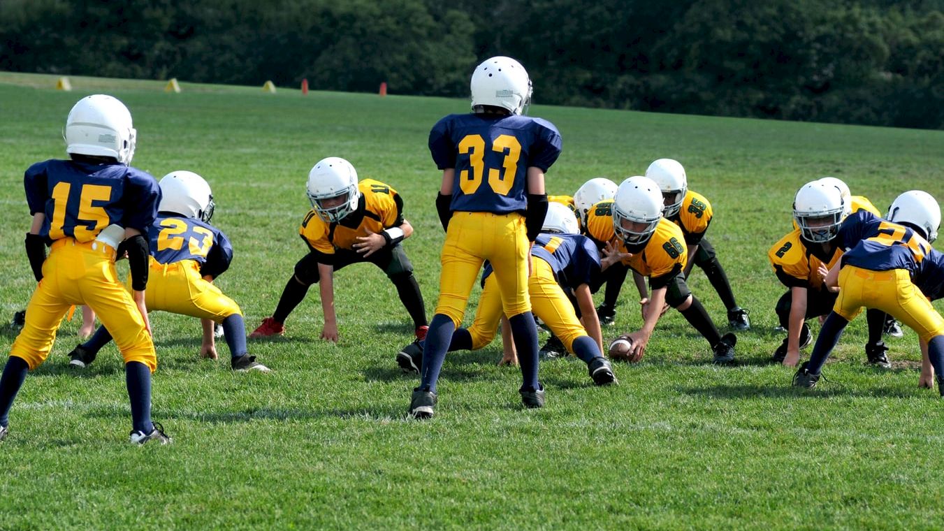 Kids wearing yellow and blue uniforms and white helmets are playing football on a grassy field, getting ready for a snap.