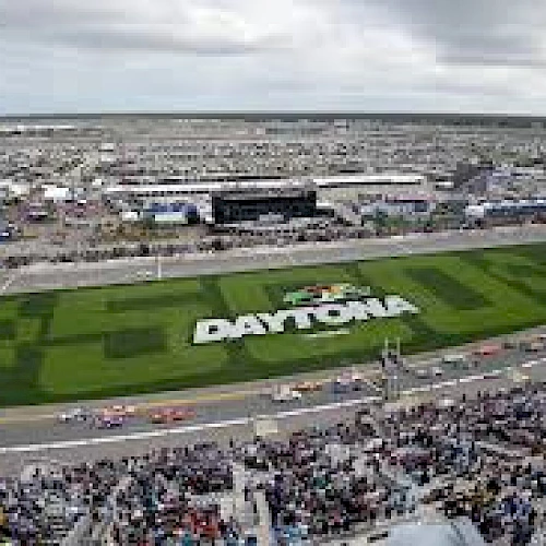 The image shows a wide view of a speedway with "Daytona" written on the grass, race cars on the track, and a large crowd in the stands.
