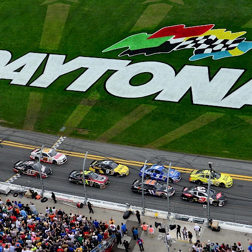 A NASCAR race at Daytona International Speedway with cars on the track, a large "Daytona" logo on the grass, and a crowd of spectators.