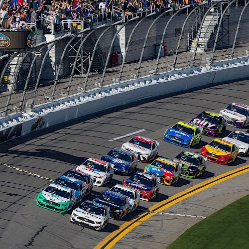 A group of race cars is on a track with a crowd of spectators in the stands, capturing an intense moment in a motorsport event.