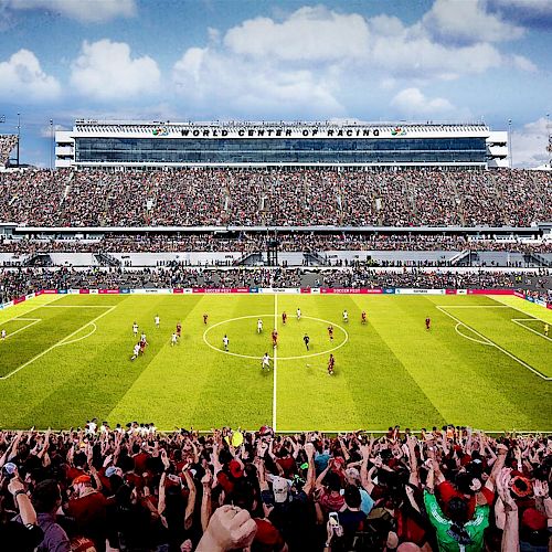 A large soccer stadium filled with spectators, with players on the field during a match. The scoreboard reads "WORLD CENTER OF RACING.".