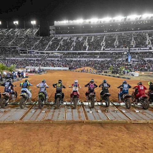 A line of motocross riders on their bikes is ready at the starting gate in a large, well-lit stadium filled with spectators at night.