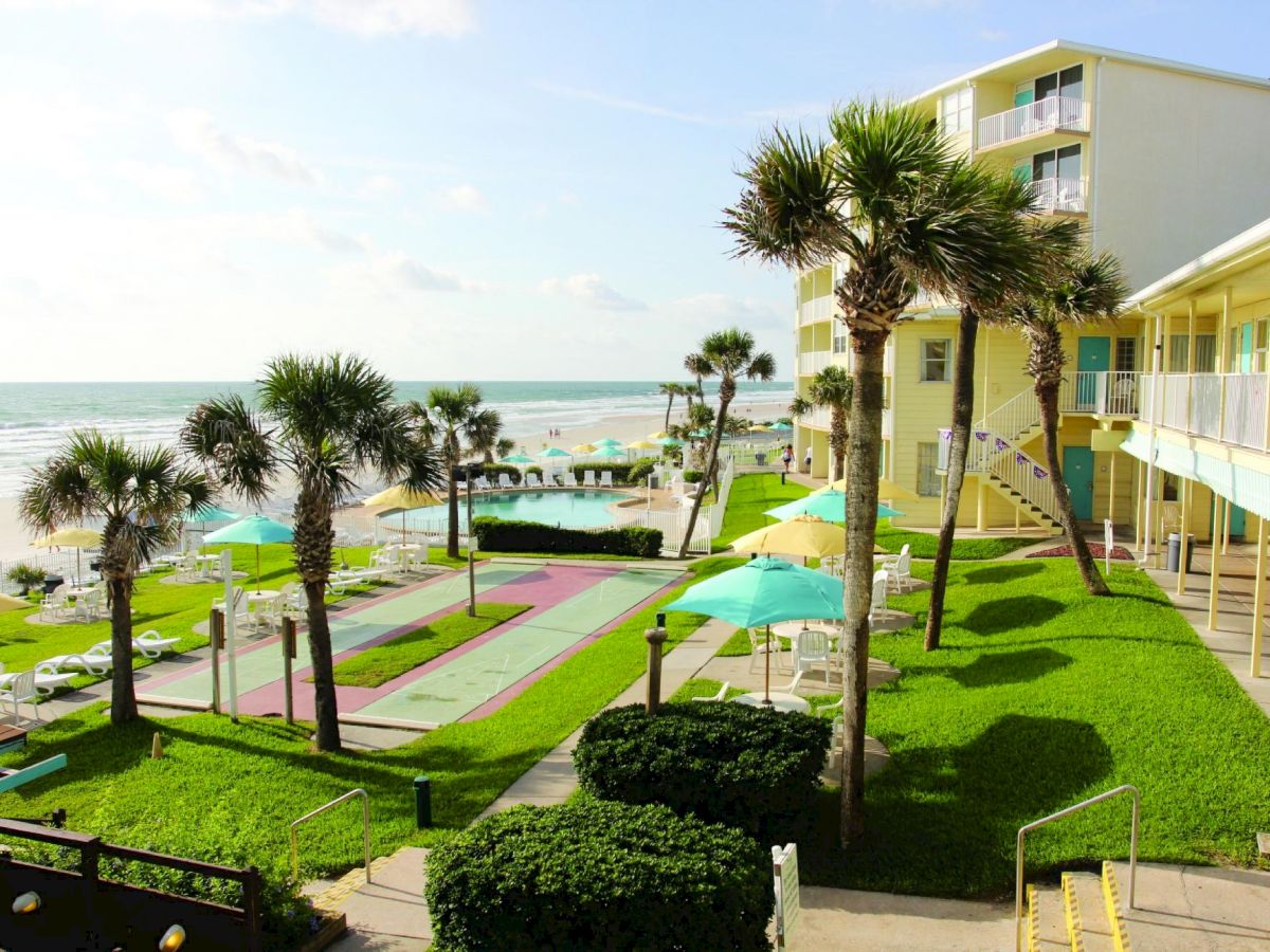 A beachfront resort with palm trees, green lawns, lounge chairs, and umbrellas set in front of the ocean, with a multistory hotel in the background.