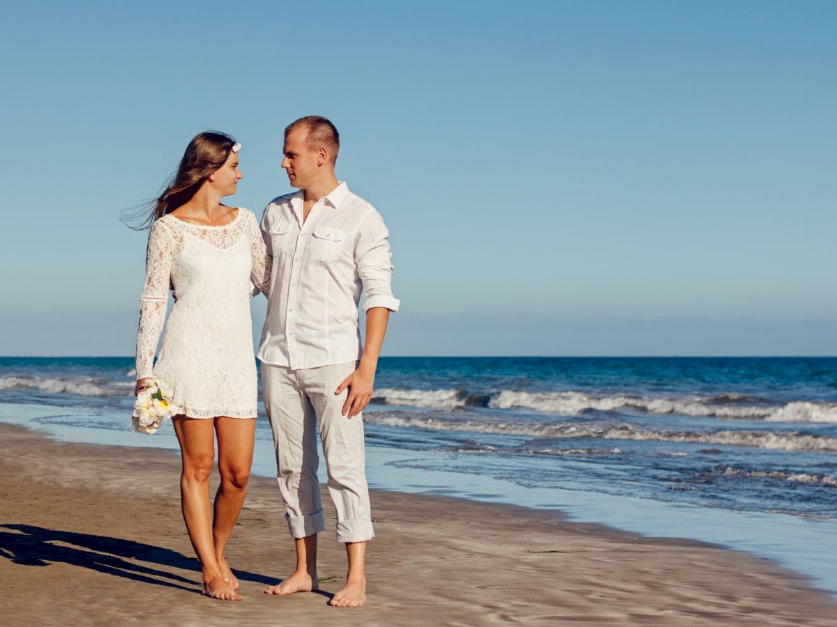 A couple, dressed in white, walks barefoot along a beach. The woman holds a small bouquet, and they share a loving gaze, with the ocean in the background.