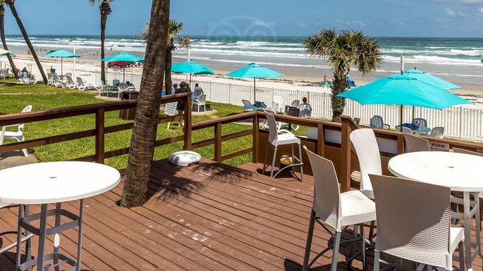 A beachside deck with tables and chairs, overlooking palm trees, umbrellas, and a sandy beach by the ocean on a bright sunny day.