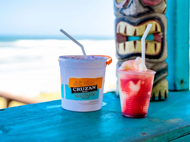 Two colorful drinks on a blue wooden table with a tiki statue and beach in the background