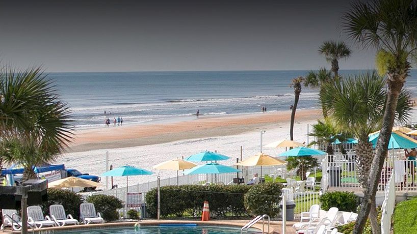 A beach scene with palm trees, a pool area with chairs and umbrellas, and the ocean in the background. A building stands to the right, ending the sentence.