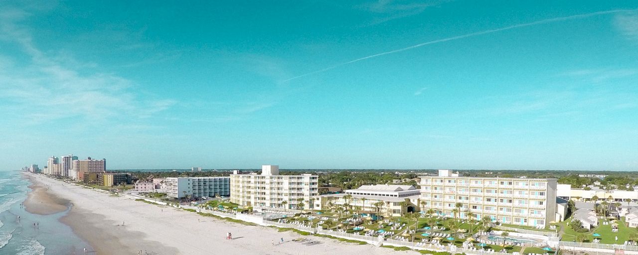 A wide aerial view of a beachfront resort with white buildings, palm trees, and a sandy beach along the ocean.