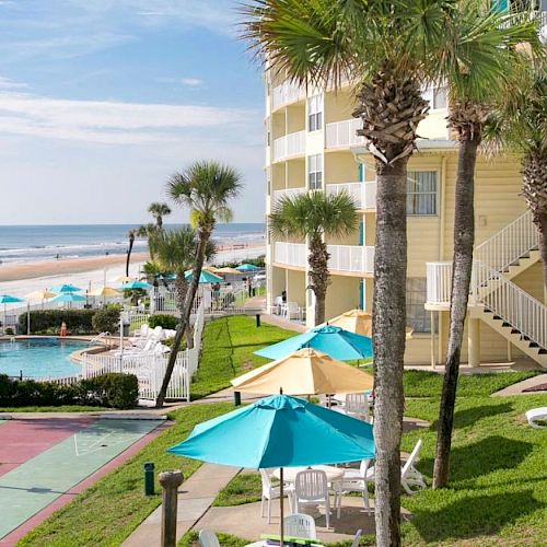 A beachfront resort features palm trees, buildings, a pool, lounge chairs, and umbrellas, with beach access and ocean views in the background.