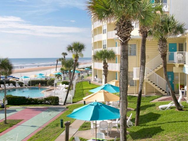 A beachfront resort exterior with palm trees, a pool, and a shuttle court in front of the ocean.