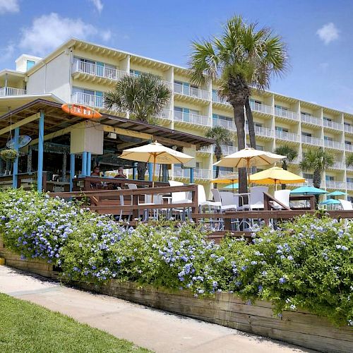 A beachfront hotel with multiple floors, outdoor seating, umbrellas, lush greenery, and palm trees against a blue sky.