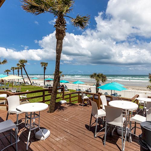 An outdoor deck with tables and chairs overlooking a beach with palm trees, lounge chairs, and blue umbrellas under a partly cloudy sky.