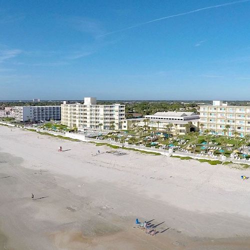 An aerial view of a beachfront with buildings and hotels lining the shore, a wide sandy beach, and a few people walking along the shoreline.