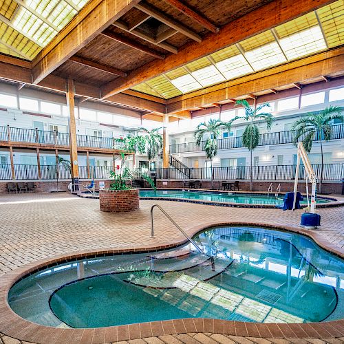 An indoor swimming facility with multiple pools, seating areas, artificial palm trees, and overhead skylights, giving a warm and inviting atmosphere.