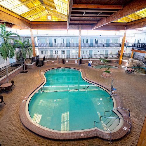 An indoor pool area with tables, chairs, and palm trees, surrounded by wooden beams and railings, under a skylight.