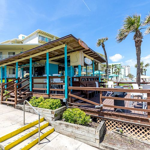 An outdoor deck with tables and chairs at a beachfront bar or restaurant, featuring palm trees and a clear blue sky in the background.