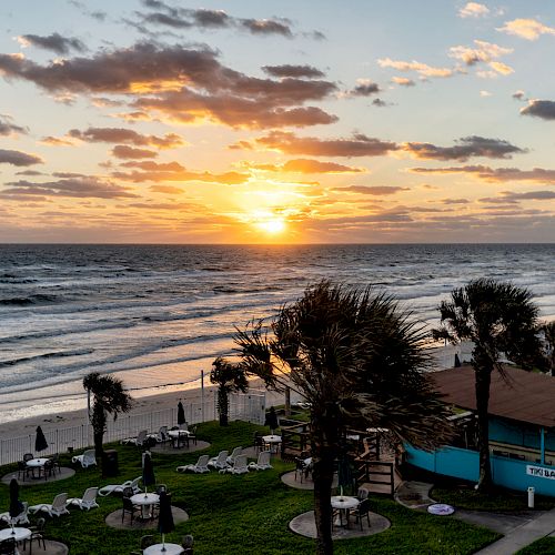 A beautiful beach scene at sunrise or sunset, with tables under umbrellas, palm trees, a small building, and waves rolling in from the ocean.