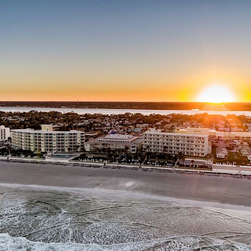 An aerial view of a beachfront with numerous buildings and a vibrant sunset on the horizon, casting a warm glow across the scene.