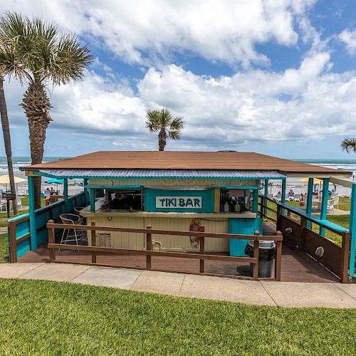 A beachside tiki bar surrounded by palm trees and a grassy area, with the ocean and beachgoers in the background.