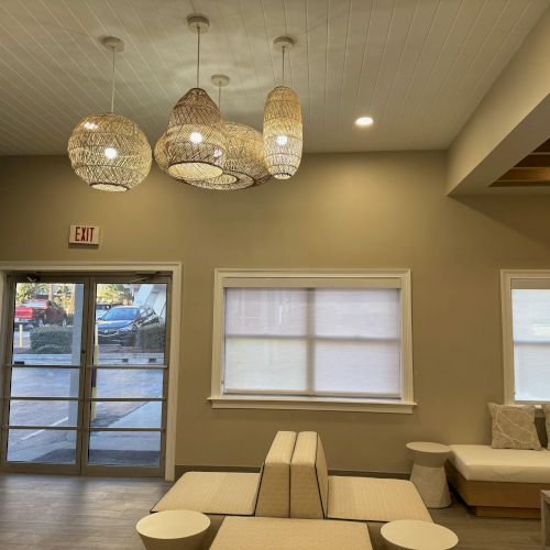 The image shows a modern indoor lounge area with wicker pendant lights, a glass exit door, window with blinds, and cushioned seats and tables.