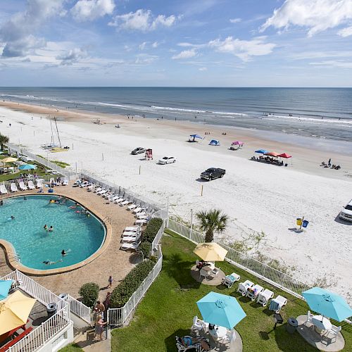 A beach scene with a swimming pool, lounge chairs, umbrellas, and people relaxing near the ocean. Cars and tents are on the sandy shore.