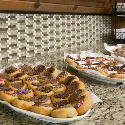 The image shows platters of assorted donuts with various toppings, displayed on a granite countertop in front of a tiled backsplash.