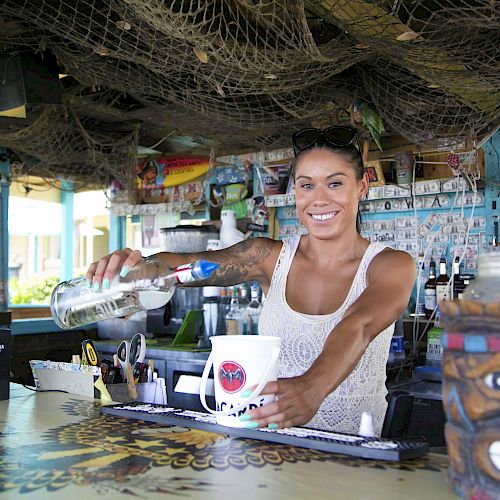 A person behind a bar pours a drink into a cup, with a tiki-themed container and hanging decor around.