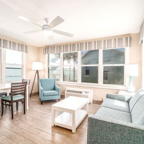 A bright living room with a ceiling fan, a sofa, armchair, coffee table, lamp, dining table with chairs, and large windows surrounded by striped drapes.