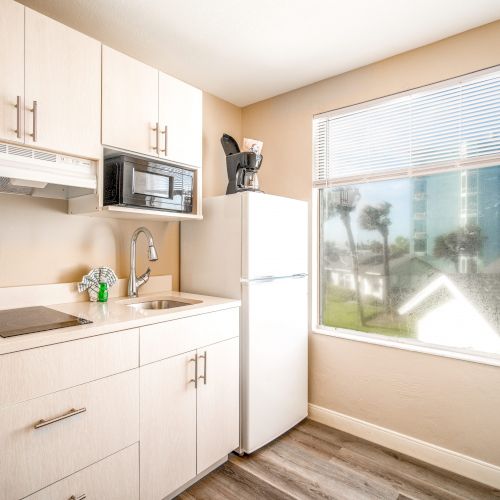 A bright kitchen with white cabinets, a sink, stove, microwave, coffee maker, and a refrigerator by a large window with blinds and outdoor view.