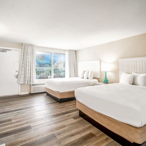 The image shows a well-lit hotel room with two double beds, white linens, a TV, a small lamp, a large window, and wooden floors, creating a cozy atmosphere.