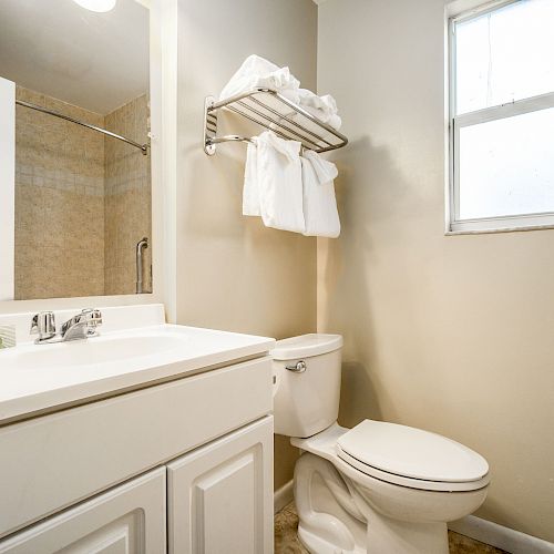 The image shows a clean bathroom with a toilet, a vanity with a sink, a wall-mounted towel rack with towels, and a window, all in light colors.