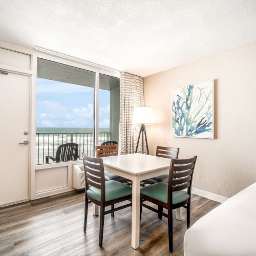 A well-lit room with a table and chairs, a bed, a large window, a painting, and a door leading to a balcony with a scenic view of the outdoors.