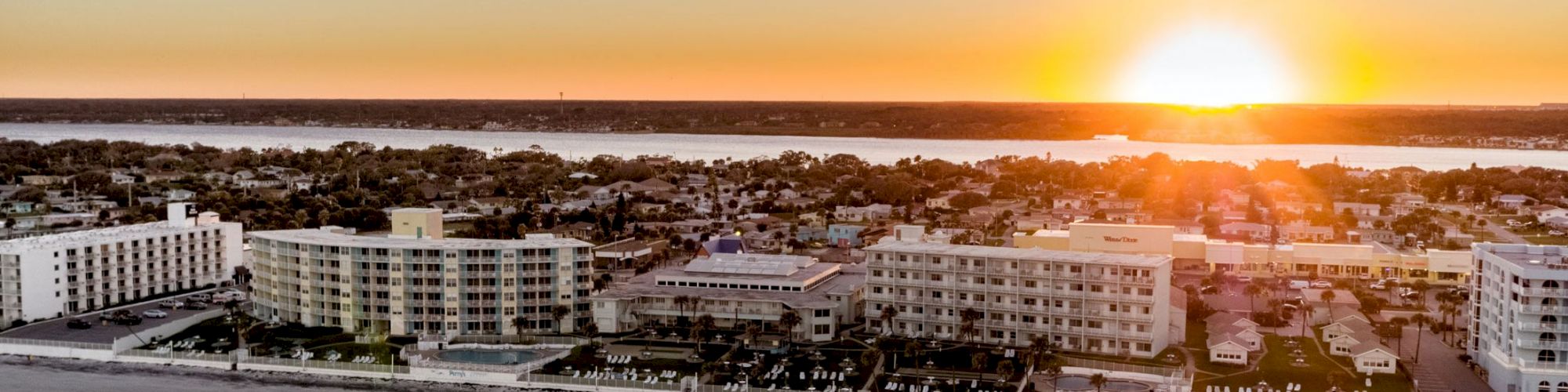 An aerial view of beachfront buildings and homes at sunset, with a clear sky and the sun setting on the horizon over a distant water body.