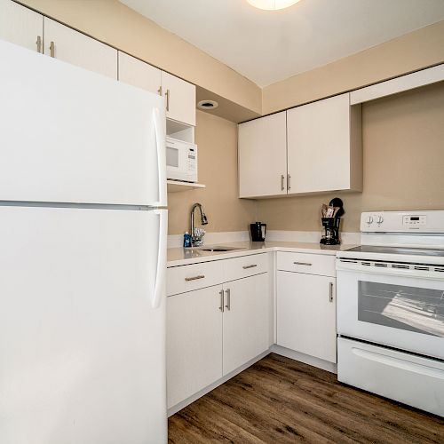 This image shows a compact kitchen with white cabinets, a refrigerator, a microwave, and a stove. The floor is made of wood.