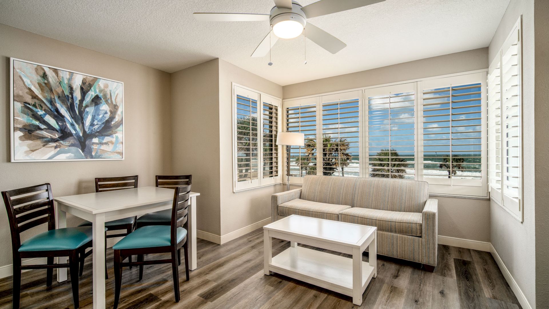 A bright living room with ocean views through large windows, featuring a sofa, coffee table, dining set, ceiling fan, painting, and wooden flooring.