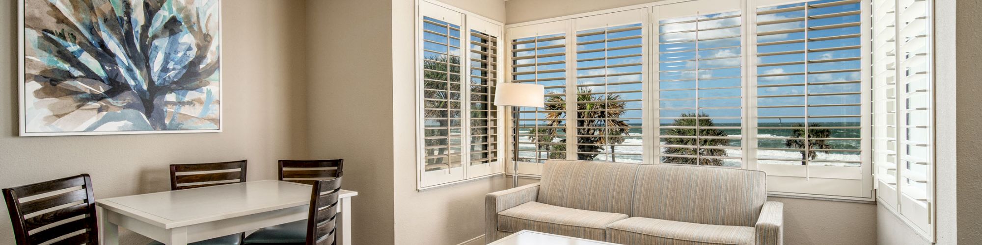 A bright living room with ocean views through large windows, featuring a sofa, coffee table, dining set, ceiling fan, painting, and wooden flooring.