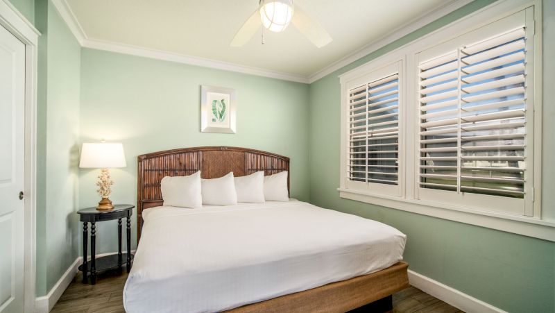 A neatly made bed with white linens and pillows in a room with light green walls, a window with blinds, and a small table with a lamp.
