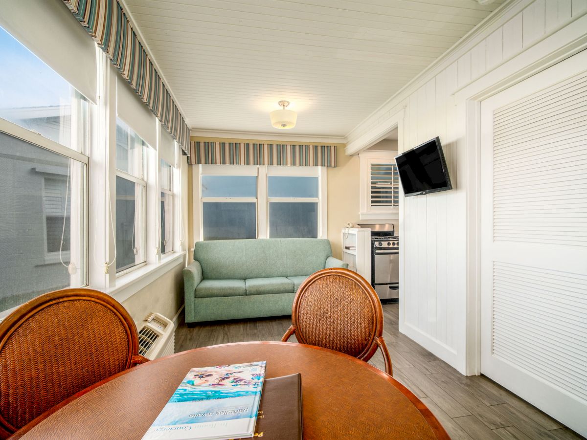 This image shows a bright, compact living space with a table, chairs, a sofa, a wall-mounted TV, and a kitchen area.