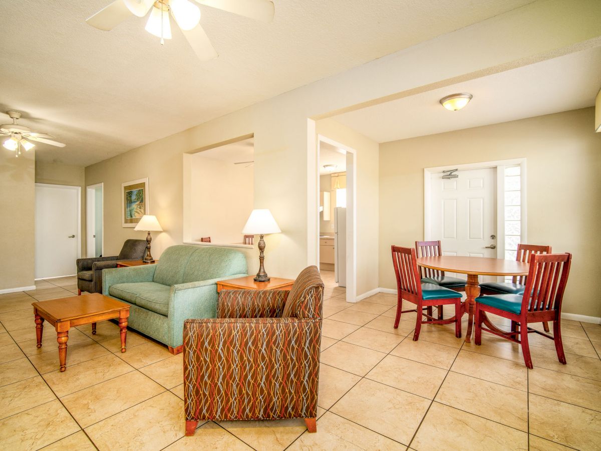 A living space with tiled floors, green and beige furniture, a dining table with red chairs, two lamps, ceiling fans, and a door in the background.
