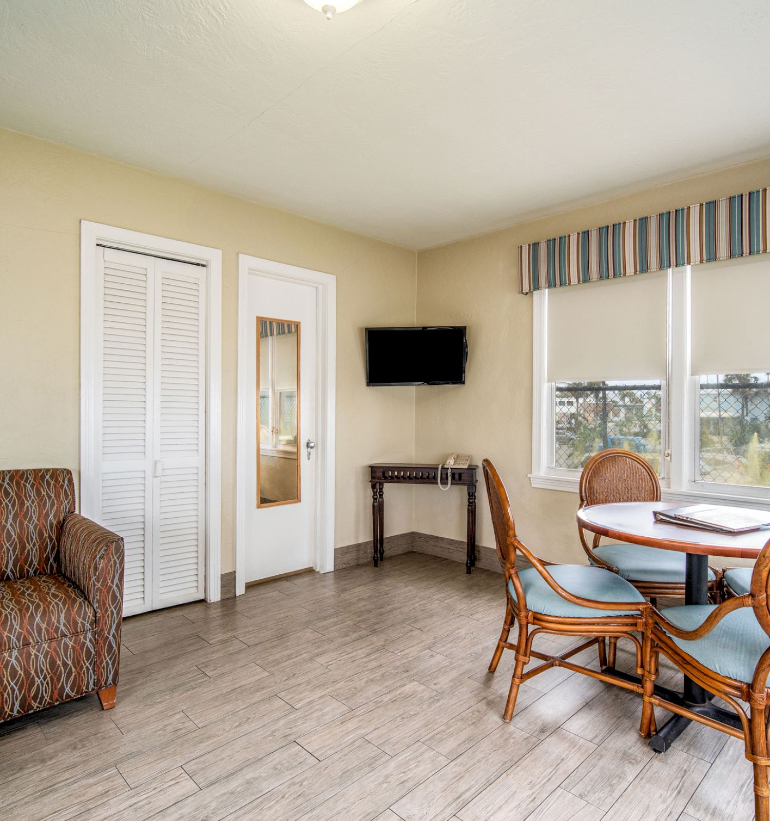 The image shows a small, cozy room with a chair, a table with four chairs, a wall-mounted TV, and a window with a striped valance.