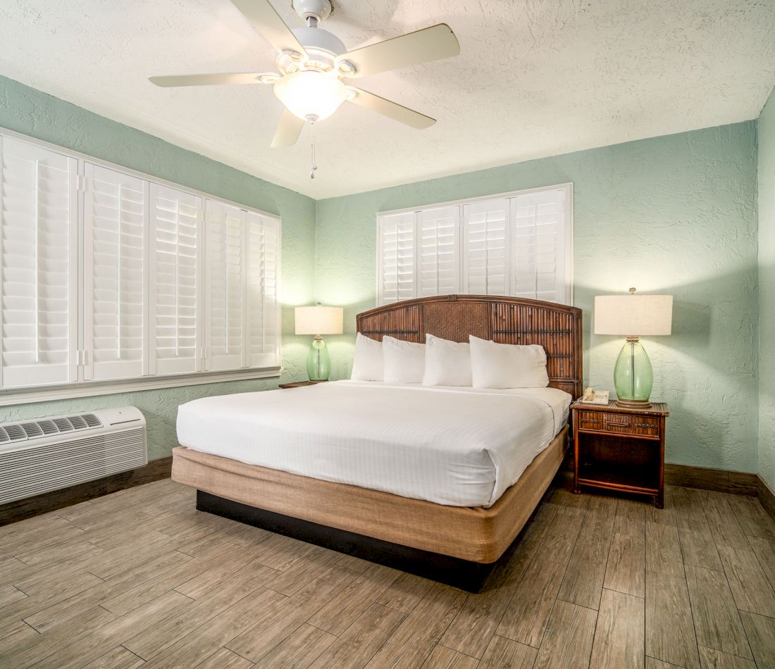 A wide-angle view of a guest room with a large bed, ceiling fan, lamps, and shutters on the windows.
