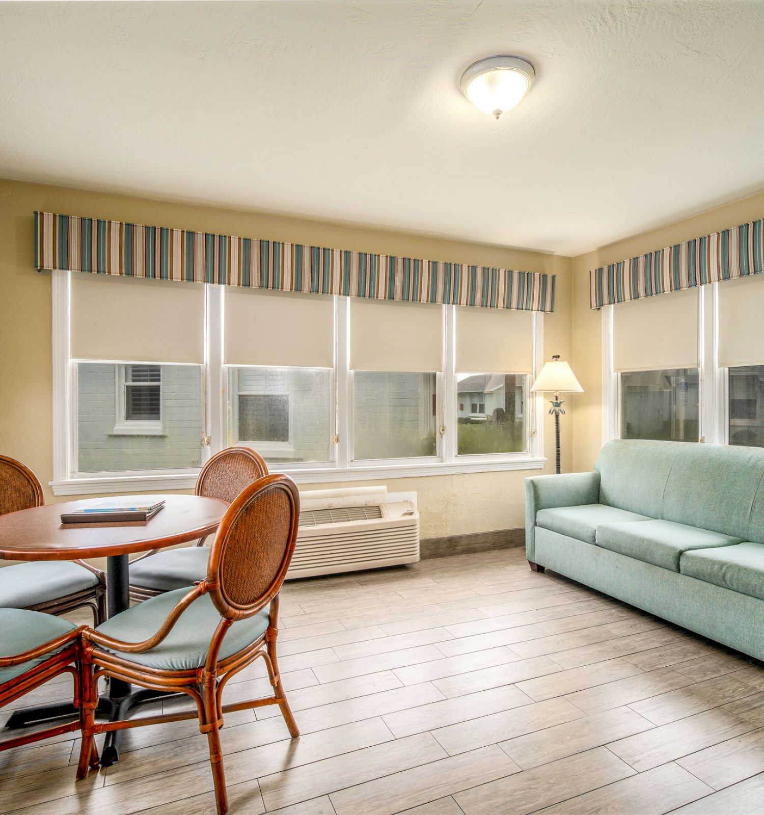 A bright room with a round dining table and wicker chairs, a green sofa, large windows with striped valances, and a floor lamp in the corner.